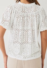 Blouse en dentelle blanc avec des manches bouffantes et des bords festonnés portée par une personne de dos, associée à un pantalon beige.