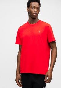 Άνδρας που φοράει φωτεινή κόκκινη κοντομάνικη μπλούζα με μικρό λευκό λογότυπο "cK" στο στήθος, συνδυασμένη με μαύρο παντελόνι, μπροστά σε απλό φόντο.