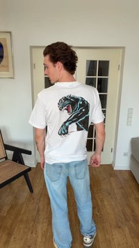 Jauns vīrietis baltā T-kreklā ar lielu rūcoša melna panteras attēlu uz muguras stāv gaišā telpā ar koka grīdu.