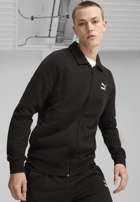 Sort zip-up jakke i blødt stof med krave og ribbede manchetter. Har et lille hvidt Puma-logo på brystet. Stylet med matchende bukser.