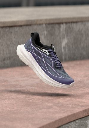 Chaussure de course athlétique violette et noire avec une semelle blanche épaisse, flottant au-dessus d'un pavé en pierre.