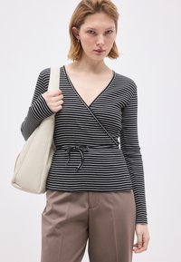 Γυναίκα που φοράει μαύρο και άσπρο ριγέ τοπ με μακριά μανίκια και μπεζ παντελόνι, κρατώντας μια μεγάλη λευκή τσάντα ώμου.