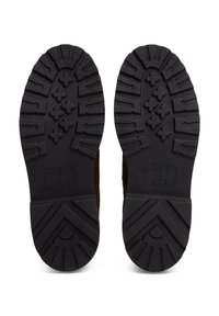 Suelas de goma de botas de color marrón oscuro con profundas ranuras y logotipo rectangular de Tommy Jeans embosado cerca del talón sobre fondo blanco.