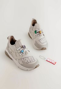 Baskets beige clair avec un dessus texturé, présentant la marque Marvel, des charmes colorés de personnages sur les lacets et un porte-clés avec un logo rouge.