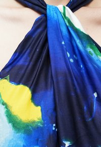 Monivärinen haltertoppi, joka on valmistettu sileästä kankaasta ja jossa on sinisiä, vihreitä ja keltaisia abstrakteja kuvioita. Niskalenkillä solmittava malli, jossa on pehmeää laskostusta.