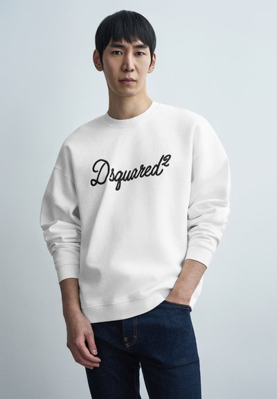 Weißes Sweatshirt mit schwarzem "Dsquared²"-Logo, Rundhalsausschnitt und gerippten Bündchen. Kombiniert mit dunklen Denim-Jeans steht das Model vor einem hellen Hintergrund.