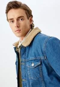 Giubbotto in denim con colletto in shearling beige, dotato di tasche sul petto e laterali, con cuciture a contrasto. Indossato sopra una camicia verde.