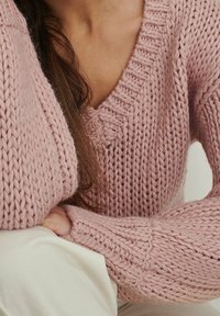 Gruby sweter z dzianiny w miękkim różowym kolorze, z głębokim dekoltem w kształcie litery V, teksturowanymi rękawami i prążkowanymi detalami na dekolcie i mankietach.