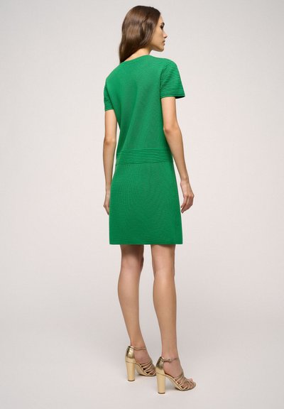 Abito verde a maglia con maniche corte, caratterizzato da strisce orizzontali testurizzate sulle spalle e in vita. Abbinato a sandali con tacco alto e cinturini dorati.