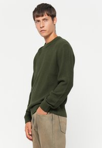 Giovane con capelli corti castani che indossa un pullover tessuto verde scuro e pantaloni marroni chiari, in piedi con una mano in tasca.