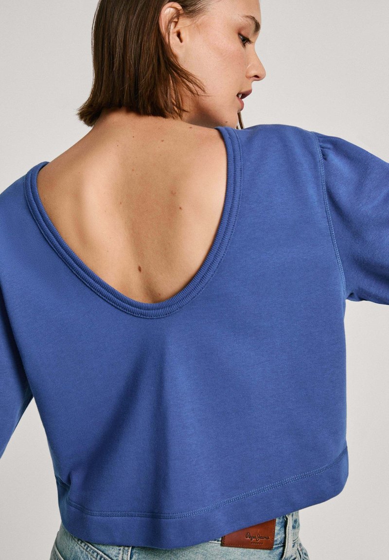 Blauwe cropped sweatshirt met een diepe V-rug, gemaakt van zachte stof. Heeft lange mouwen en zichtbare stikseldetails aan de zoom en de halslijn.