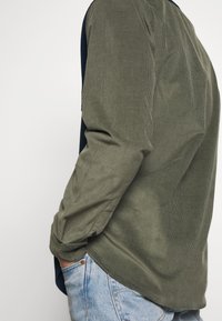 Vähän pitkähihainen paita tummanvihreällä resorakenteella, yhdistetty siniseen denimiin. Siinä on kaareva helma ja napitettavat kalvosimet.