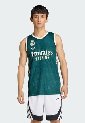 Młody mężczyzna ubrany w zieloną koszulkę do koszykówki Real Madrid z tekstem "Emirates Fly Better" oraz białe sportowe spodenki Adidas na jednolitym tle.