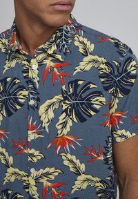 Korte mouwen overhemd in blauwe stof met een tropisch blad- en bloemenpatroon in groen, rood en geel. Heeft een puntige kraag en knopen.
