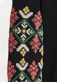 Czarna tkanina z haftowanymi wzorami geomtrycznymi i kwiatowymi w kolorach zielonym, żółtym, białym i czerwonym, ułożonymi w symetryczne kształty rombów.