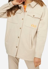 Veste beige oversize à boutons, confectionnée en tissu doux, dotée de deux poches poitrine, d'une poche latérale et d'une broderie représentant un symbole infini orange.