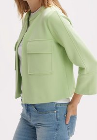 Veste courte vert clair à encolure ronde avec deux poches avant, assortie à un jean bleu ajusté. Texture lisse et coupe décontractée.