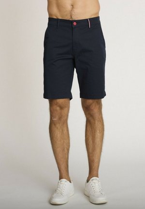 Homme portant un short bleu marine mi-genou avec un bouton rouge et des baskets blanches, debout devant un fond uni.