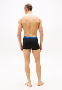 Boxer shorts negros hechos de material elástico con una cinturilla azul y roja que lleva el nombre de la marca. Vista desde la parte de atrás, sin detalles sobre el tono de piel.