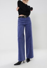 Vrouw in hoogwaist wijde paarse broek, zwarte spitse hakken, cropped zwart truitje en zwarte riem met hartvormige gesp.