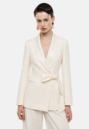 Donna che indossa un blazer crema con cintura annodata e pantaloni abbinati, abbinato a grandi orecchini pendenti rosa e neri.