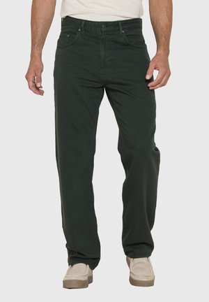 Uomo con jeans neri a gamba dritta e scarpe beige, in piedi con le mani rilassate lungo i fianchi su uno sfondo semplice.