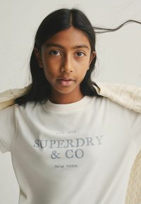Balta kokvilnas T-kreklu ar "SUPERDRY & CO" izdrukātu pelēkā krāsā. Apaļš kakla izgriezums. Gaiši dzijas krēmkrāsas džemperis novilkts pār kreiso plecu.