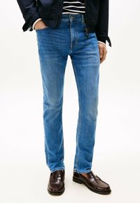 Hombre vistiendo jeans azules, mocasines de cuero marrón, una chaqueta negra y una camisa a rayas con las manos en los bolsillos, de pie sobre un fondo blanco.