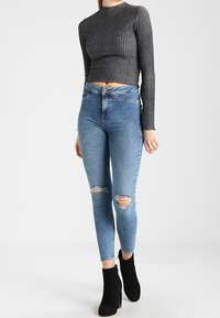 Top negro de canalé de manga larga combinado con jeans ajustados de color azul claro que presentan desgastes en las rodillas y botas negras de tacón alto hasta el tobillo.