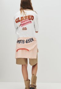 Weißes, übergroßes T-Shirt mit dem Schriftzug "GLAZED" und einer Eisschilldung auf der Rückseite. Orangegestreiftes Shirt um die Taille gebunden. Braune Shorts und Stiefel.