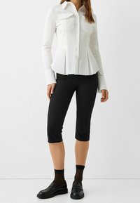 Woman wearing a fitted white pleated button-up shirt, black cropped pants, sheer black ankle socks, and black loafers, standing against a plain background.