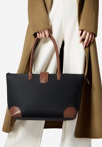Musta nailon-tote laukku ruskeilla nahkaosilla, suorakulmainen muoto, kaksi yläkahvaa ja pieni logopatch etupuolella.