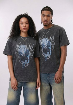 Zwei Erwachsene tragen schwarze T-Shirts mit blauen Anime-Designs und verblasste blaue Jeans und stehen nebeneinander vor einem schlichten Hintergrund.