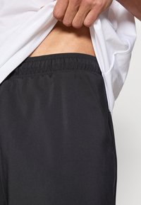 Fekete sportos rövidnadrág rugalmas derékpánttal, könnyű anyagból és oldalzsebekkel, részben egy fehér póló alatt látható.