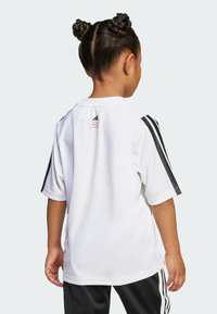 Biała sportowa koszulka z czarnymi paskami na rękawach, żebrowanym kołnierzykiem oraz logiem Adidas na plecach. Gładka faktura materiału.