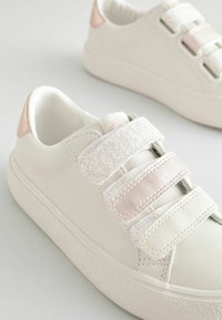 Baskets blanches avec des semelles en caoutchouc texturé, comportant trois sangles : une scintillante, une en satin rose et une lisse blanche. Accentuées par des languettes de talon roses.