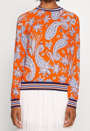 Femme portant un haut à manches longues orange vif avec des motifs floraux paisley bleus et blancs et des poignets et un ourlet rayés.
