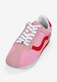 Zapatilla deportiva rosa con una franja lateral roja, cordones blancos, cuello acolchado y suela texturizada. Los materiales incluyen sintético y tela.