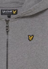 Grå zip-up hoodie tillverkad av en bomullsblandning. Har en gul broderad logotyp på bröstet och en metallisk dragkedja.