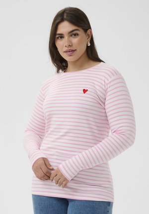 Moteris dėvi ilgomis rankovėmis dryžuotą rožinės ir baltos spalvų palaidinę su mažyte raudona širdute ant krūtinės, priderintą prie mėlynų džinsų.