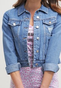 Chaqueta de mezclilla de color azul claro, diseño corto con botones de plata, dos bolsillos en el pecho, detalle desgastado en el bolsillo derecho, mangas arremangadas.