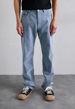 Person wearing loose light blue jeans and black canvas sneakers with tan rubber soles, standing on a gray floor against a white wall.