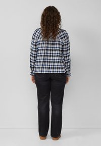 Frau mit lockigem Haar, trägt ein blau-weiß kariertes Hemd, schwarze Jeans und braune Schuhe, steht vor einer schlichten grauen Wand.