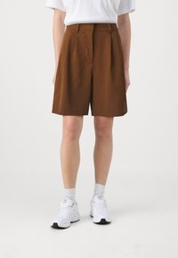 Person som bär bruna knä långa shorts, vita sportstrumpor och vita sneakers, stående mot en platt ljus bakgrund.