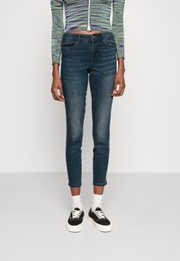 Jeans skinny de color azul oscuro con un dobladillo ligeramente desgastado, fabricados en denim. Llevados con zapatillas negras y calcetines blancos. Blusa de manga larga a rayas.