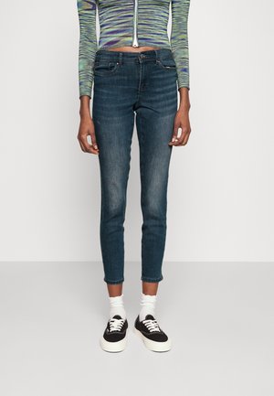 Jeans skinny de color azul oscuro con un dobladillo ligeramente desgastado, fabricados en denim. Llevados con zapatillas negras y calcetines blancos. Blusa de manga larga a rayas.
