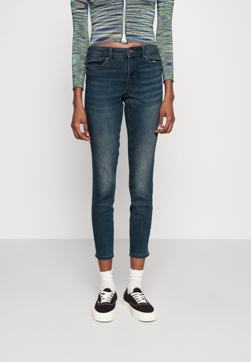 Jeans skinny de color azul oscuro con un dobladillo ligeramente desgastado, fabricados en denim. Llevados con zapatillas negras y calcetines blancos. Blusa de manga larga a rayas.