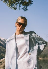 Vaaleansininen pellavapuku valkoisen paidan päällä, käytössä kilpikonnakuosiset aurinkolasit ja hopeiset renkaat; luonnollinen tausta vuorten kanssa.