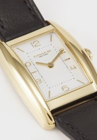 Montre rectangulaire dorée avec un cadran blanc, bracelet en cuir noir et index horaires dorés. Dispose de deux aiguilles dorées et d'un détail de couronne.