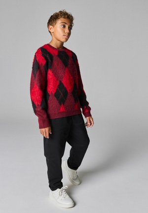 Maglione a maglia con motivo argyle rosso e nero in tessuto testurizzato, abbinato a pantaloni neri larghi e sneaker bianche.